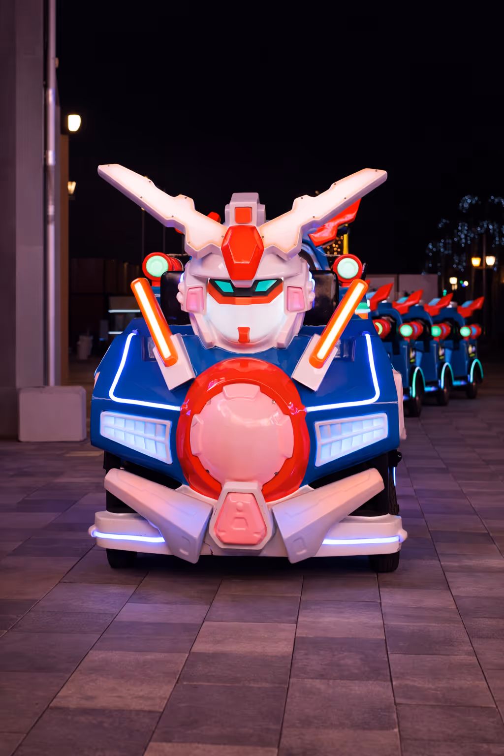 Front view of a robot-themed ride vehicle illuminated with blue and red lights lined up outdoors at night.