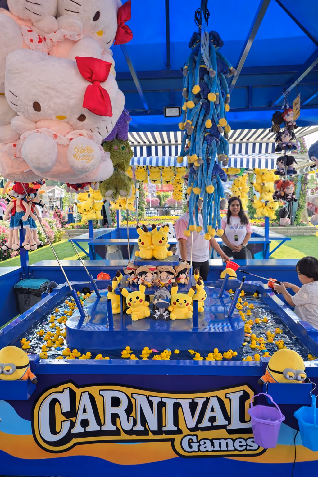 Carnival fishing game booth with hanging plush toys including Hello Kitty, Pikachu, and Minions, and a child playing.