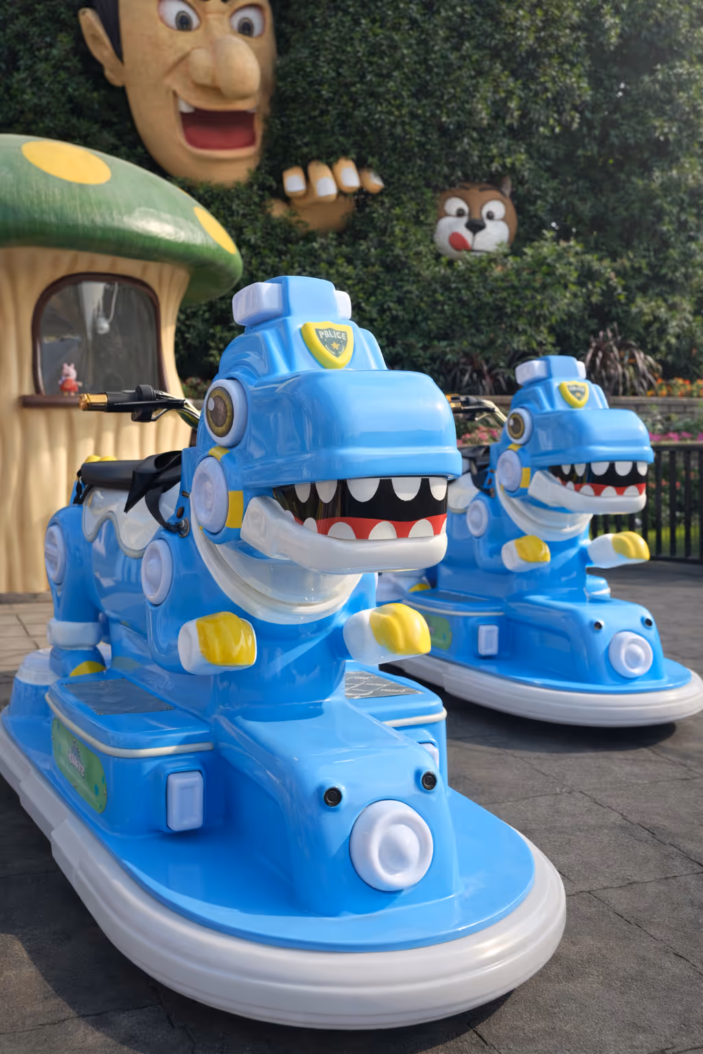 Two blue dinosaur-themed kid ride vehicles with police badges, positioned on a paved surface in a playful park setting.