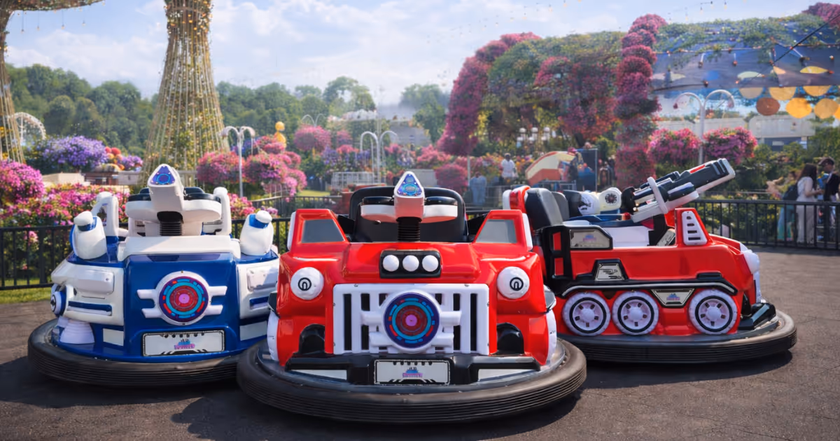Three colorful bumper cars, two red and one blue, parked in an outdoor amusement park with flowers and large tree sculptures in the background.