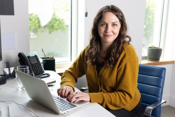 A women in an office sitting at a computer, smiling at the camera. 