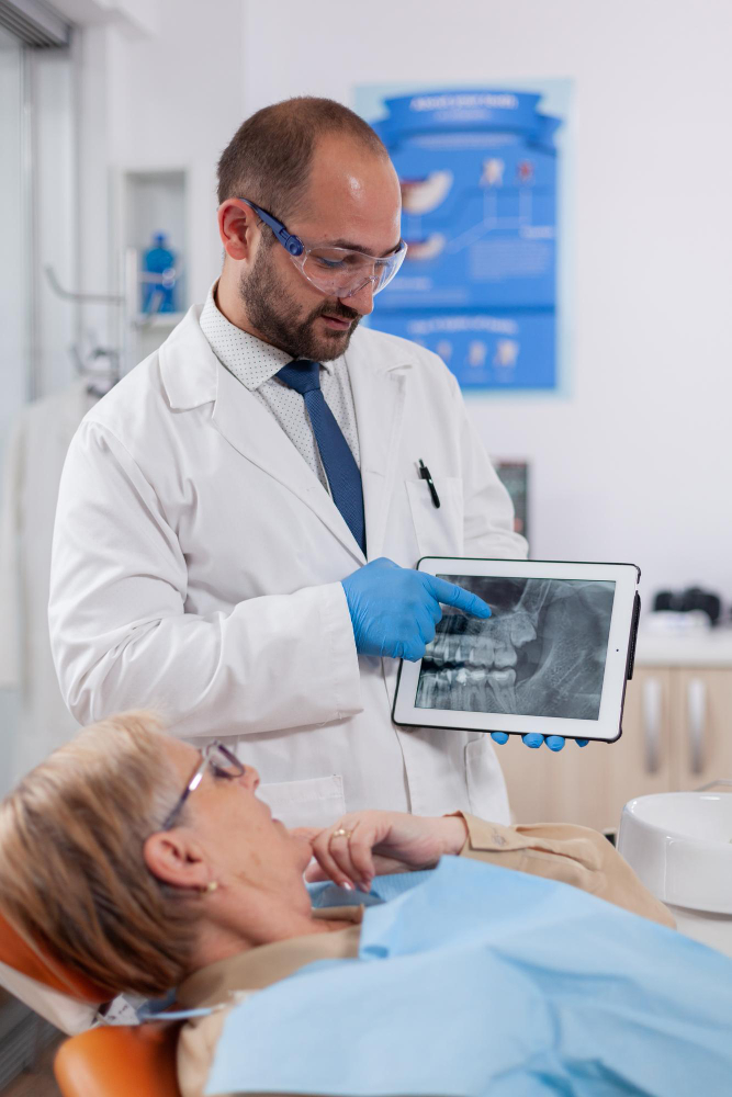 Dentist in white coat and gloves showing a dental X-ray on a tablet to a female patient reclining in a chair.