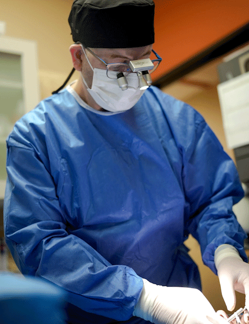 Surgeon in blue scrubs, mask, and magnifying glasses performing a surgical procedure in an operating room.