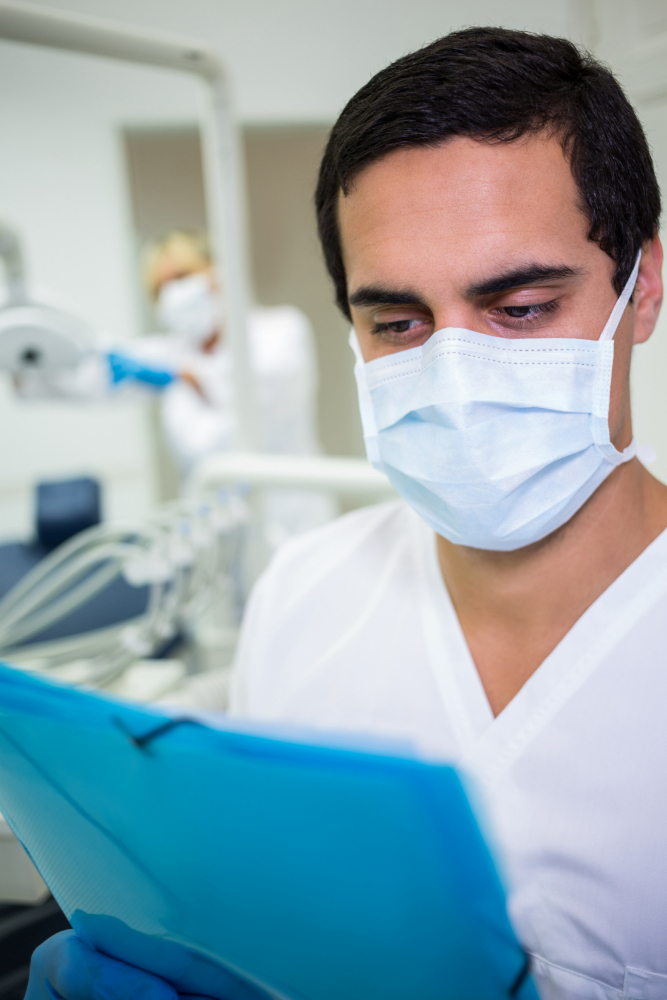Male healthcare worker wearing surgical mask and gloves, reading a blue folder in a clinic.