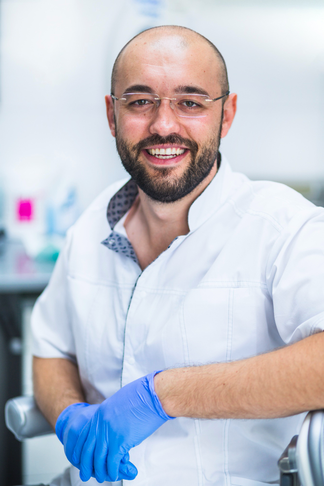 Smiling male healthcare professional wearing glasses, white coat, and blue gloves in medical setting.