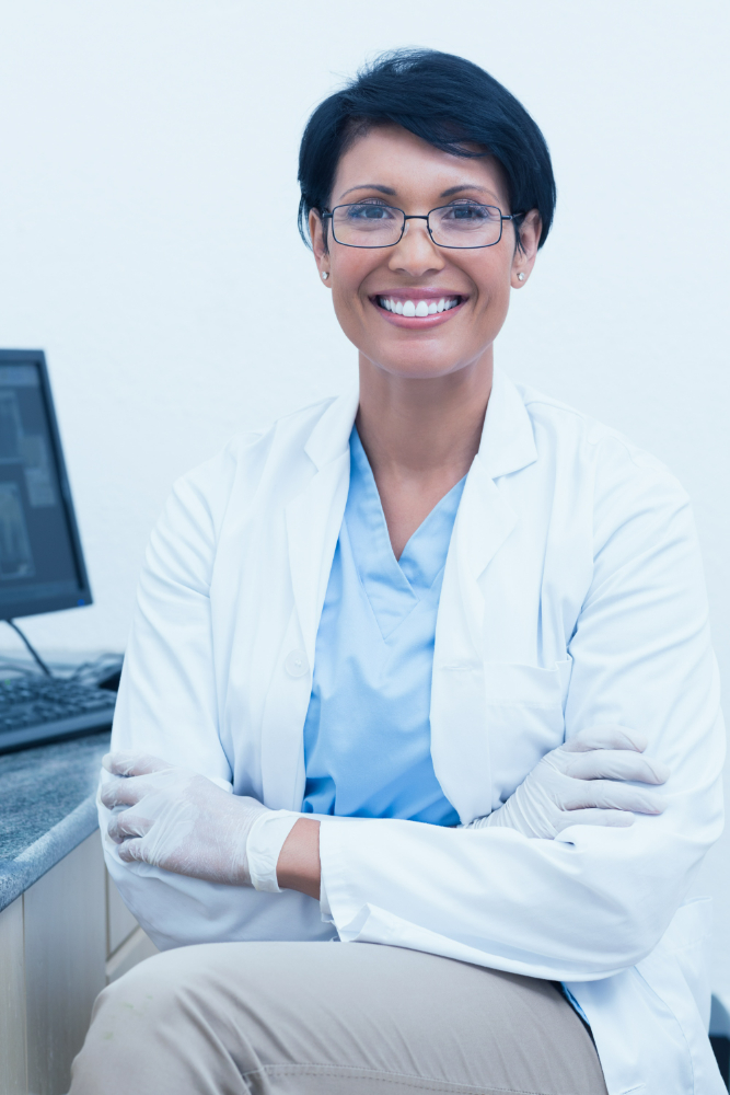Smiling female doctor wearing glasses, white coat, and gloves seated near computer in medical office.