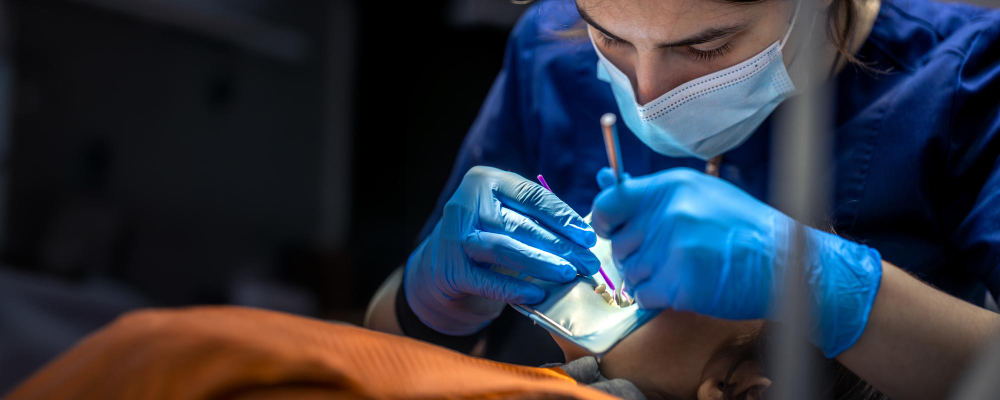 Dentist wearing mask and gloves working on a patient's teeth using dental tools and mirror.