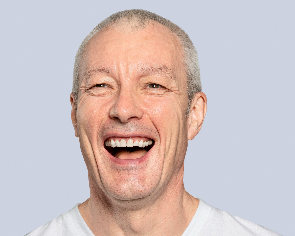 Smiling middle-aged man with short gray hair wearing a white shirt on a light gray background.