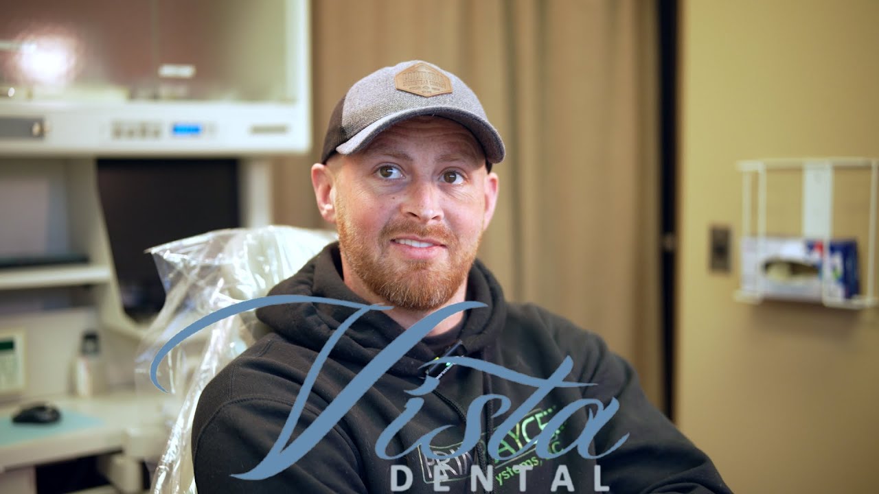 Smiling man with beard wearing a cap and black hoodie sitting in a dental office chair.