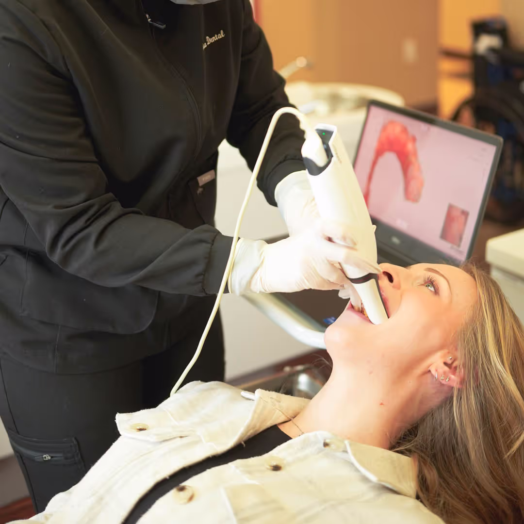 Dental professional using a scanning device inside a patient's mouth with a 3D dental model on a laptop screen.