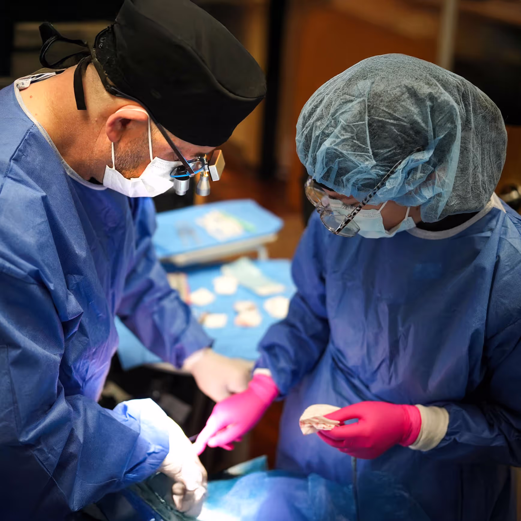 Two medical professionals in blue gowns and masks performing surgery under bright light.