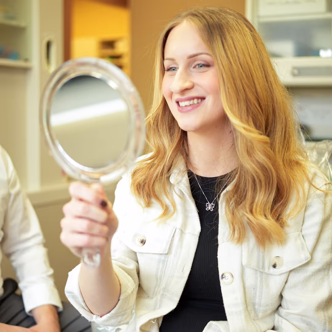 Smiling woman with long blonde hair holding a round hand mirror indoors.