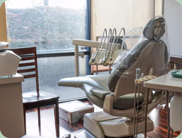 Dental chair covered in plastic next to a wooden chair by a window in a clean dental office.