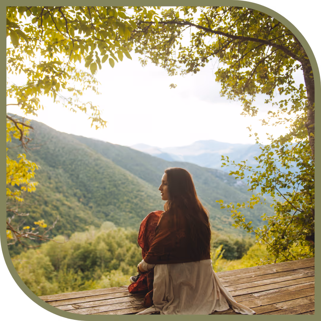 Woman with long hair sitting on wooden platform, overlooking green mountains framed by tree branches.