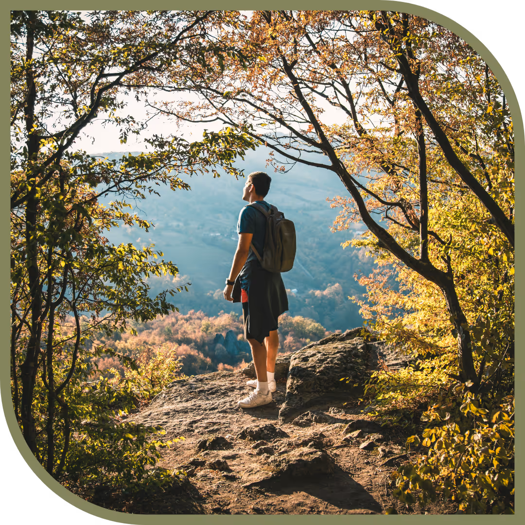 A man wearing a backpack stands on a rocky cliff surrounded by trees with autumn leaves, overlooking a valley and distant hills.