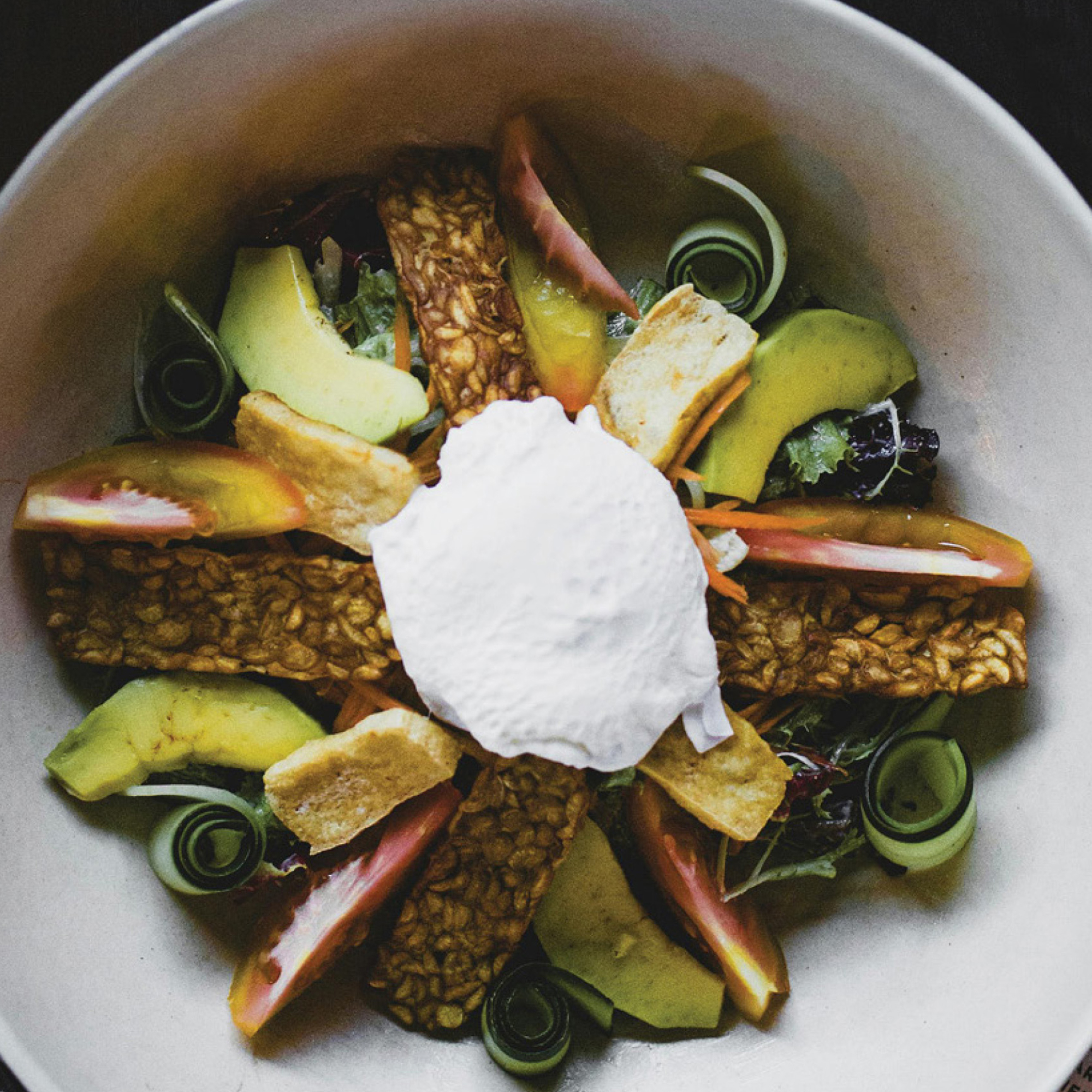 Vegan salad bowl with tempeh, avocado, heirloom tomato, cucumber spirals, mixed greens, and coconut cream topping.