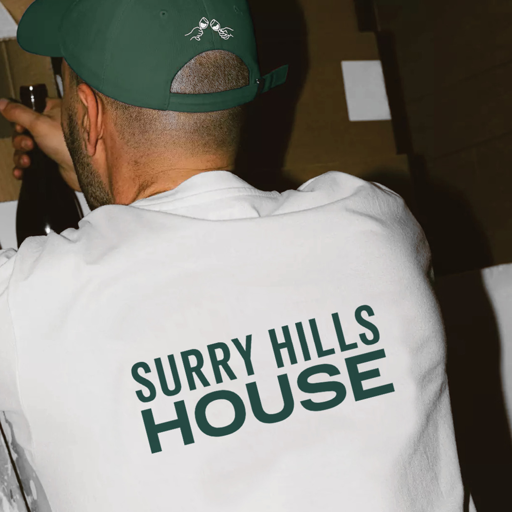 Man wearing green cap and white t-shirt with Surry Hills House logo, pouring wine indoors.