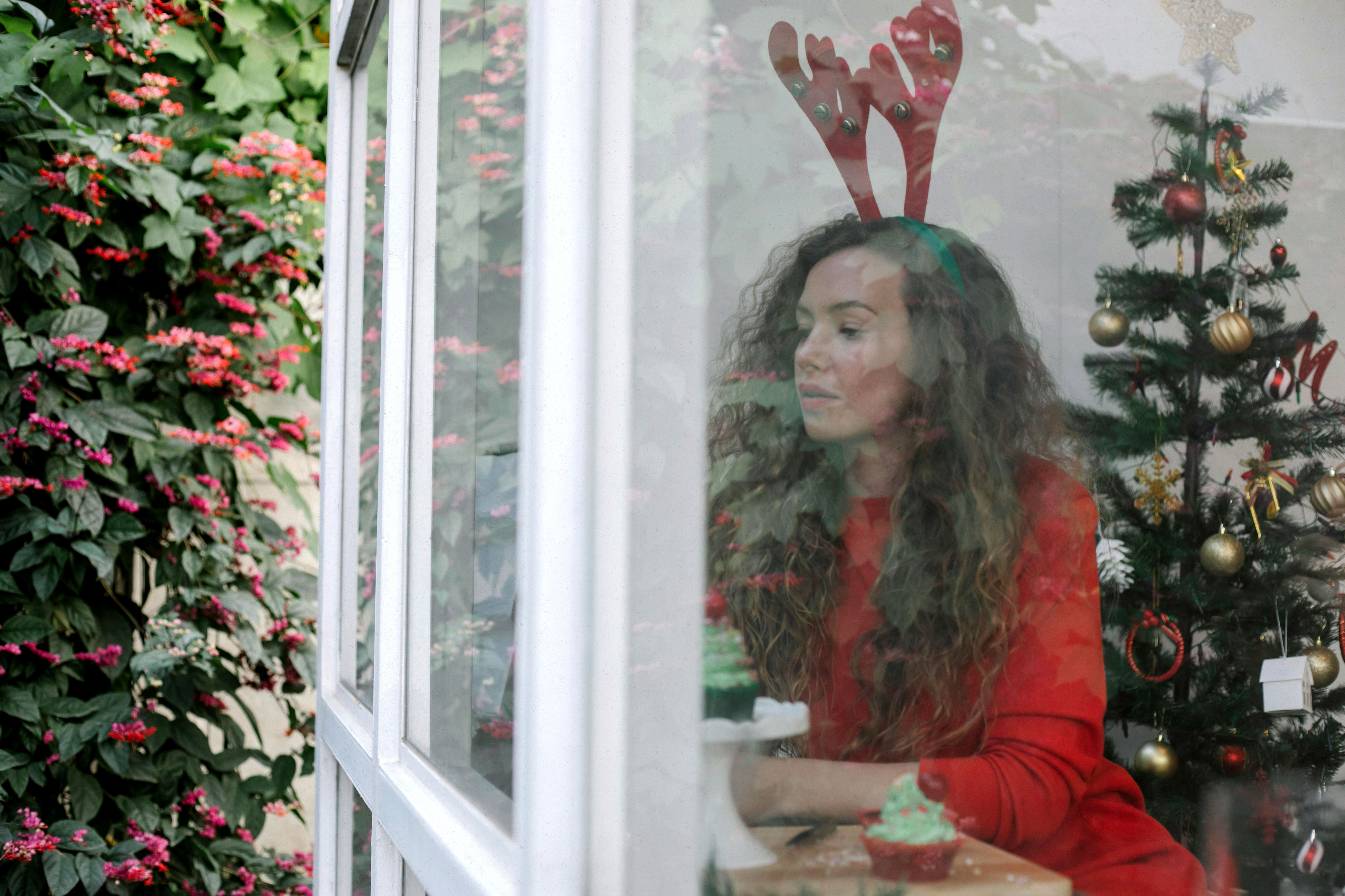 Woman sitting alone by a window during the holidays, wearing reindeer antlers and looking reflective, illustrating grief and loneliness after a breakup or divorce during the holiday season.