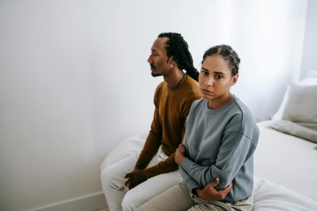 Couple sitting apart on bed with visible tension, woman looking concerned about possible infidelity