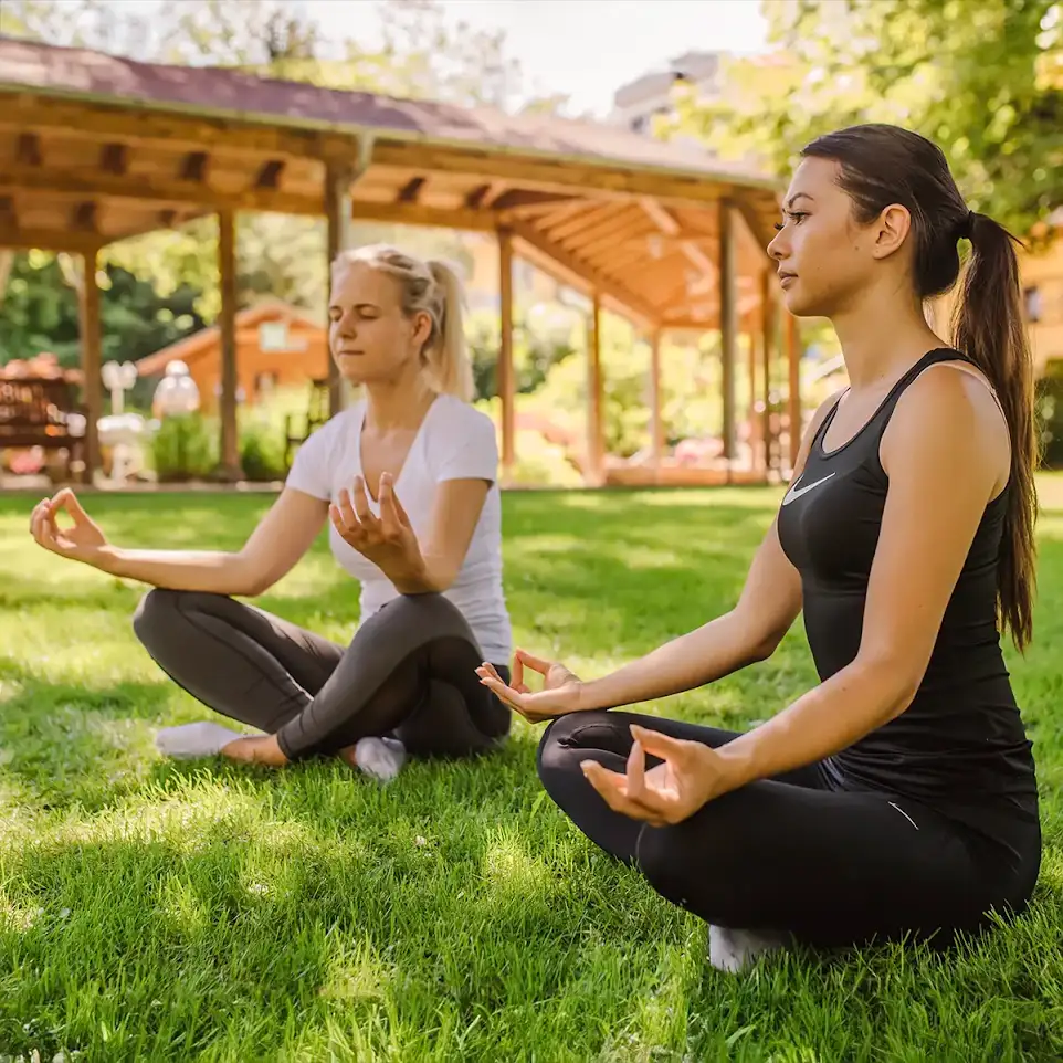 Zwei Frauen sitzen im Schneidersitz im Gras und meditieren.