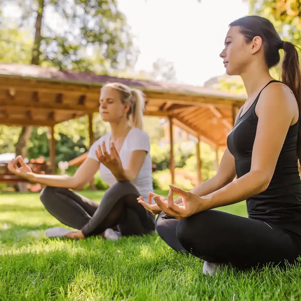 Zwei Frauen sitzen im Gras im Schneidersitz bei einer Atemübung.