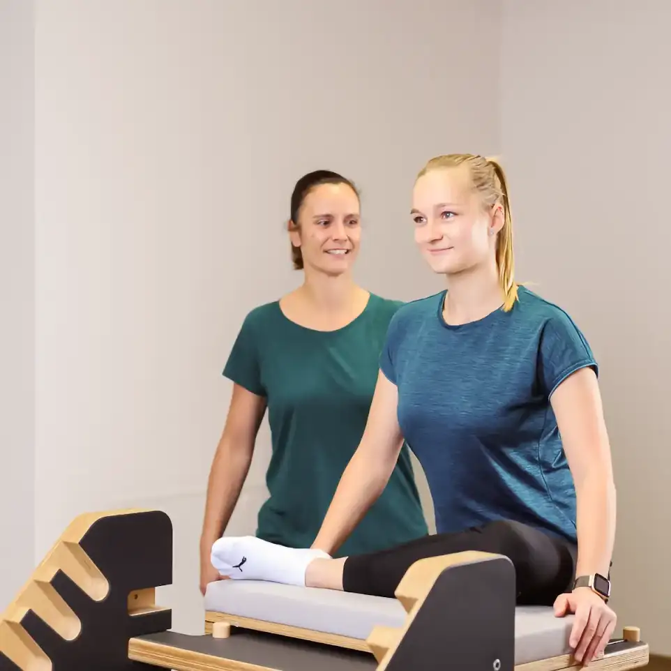 Eine Patientin sitzt mit angewinkeltem Bein auf einem Polster. Hinter ihr steht eine Therapeutin.