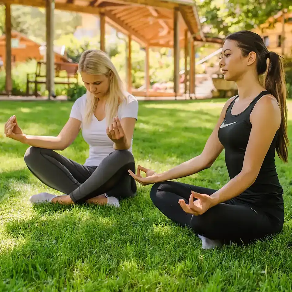 Zwei Frauen sitzen im Gras im Schneidersitz und meditieren.