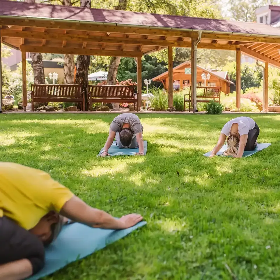 Mehrere Patienten machen draußen gemeinsam Yoga. Sie liegen vornübergebeugt auf den Yogamatten.