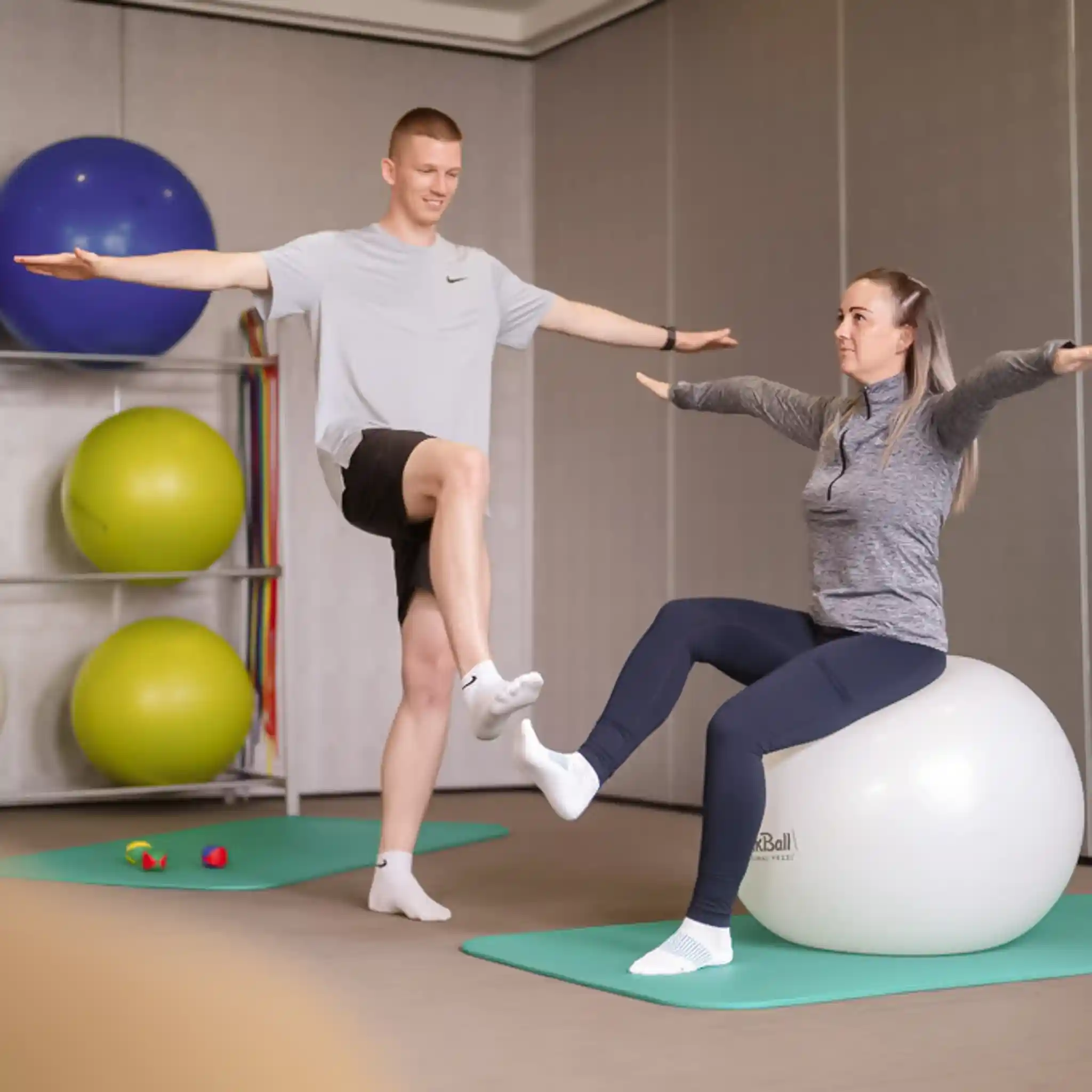 Eine Patientin sitzt auf eine Gymnasikball und macht eine Übung. Neben ihr steht ein Patient, der ebenfalls eine Übung macht.