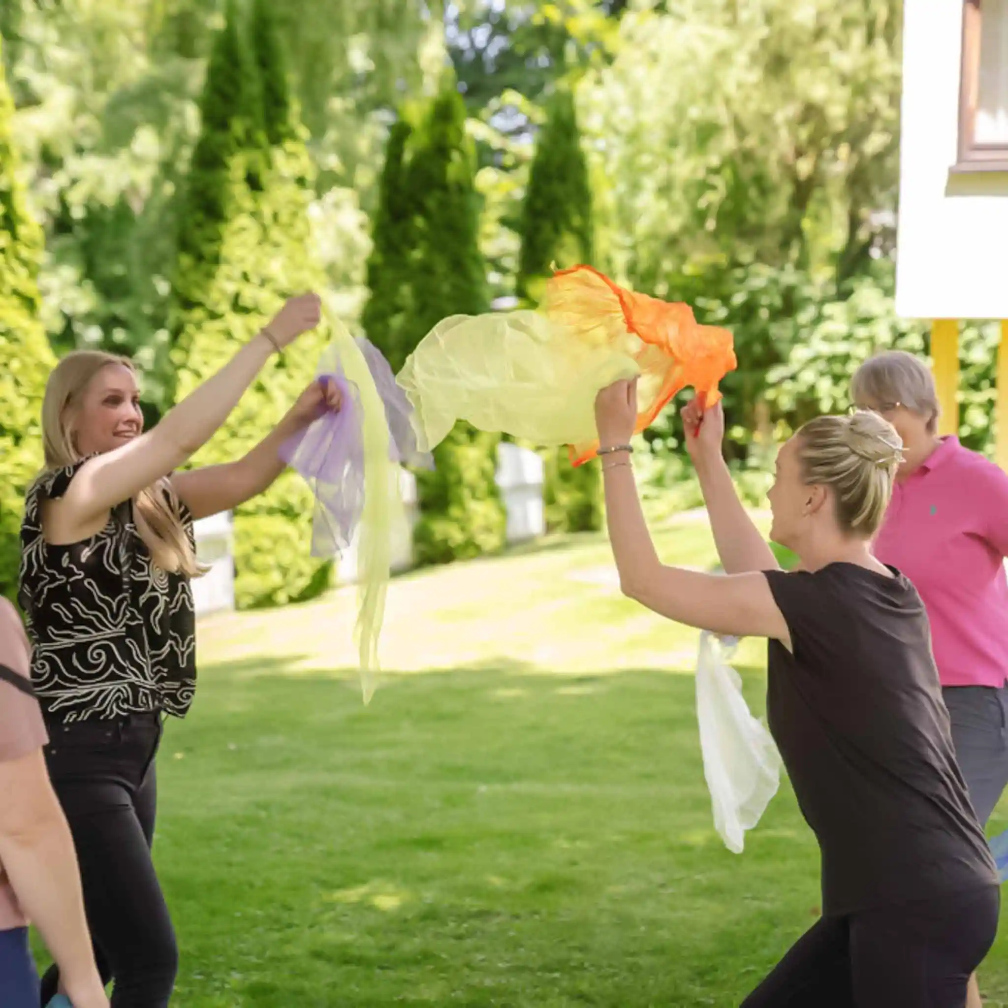 Drei Frauen wirbeln im Garten bunte Tücher in die Luft bei einer Tanztherapie.