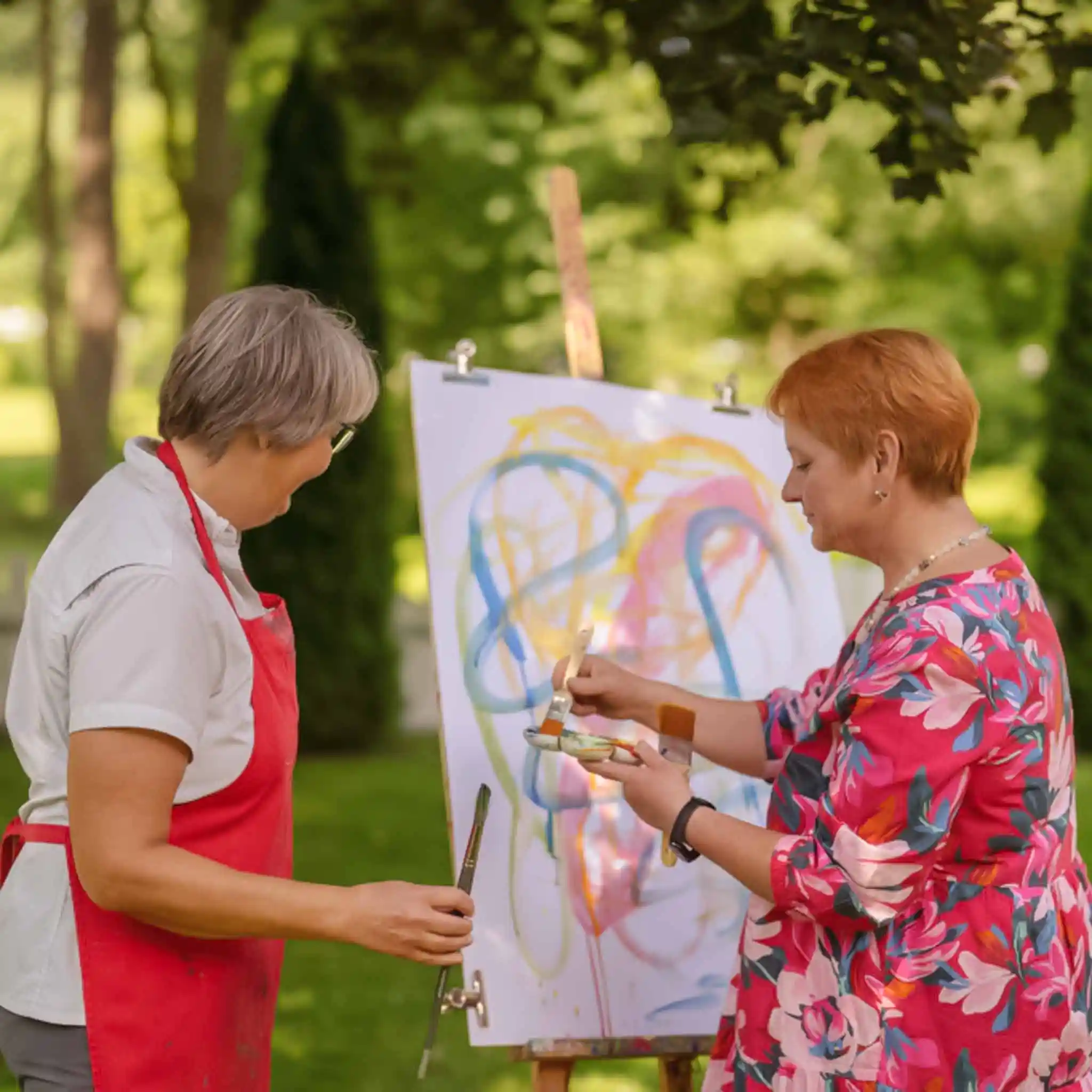 Eine Patientin malt im Garten auf einer Leinwand. Neben ihr steht eine Therapeutin.
