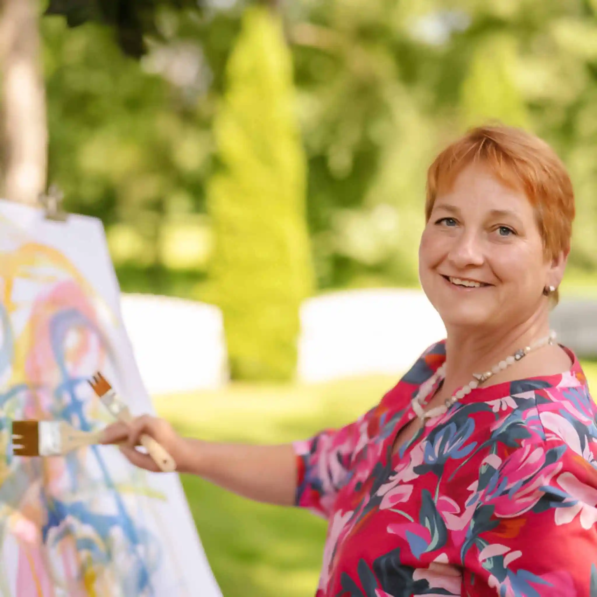 Eine Patientin malt im Rahmen der Kunsttherapie auf einer Leinwand im Garten und lacht in die Kamera.