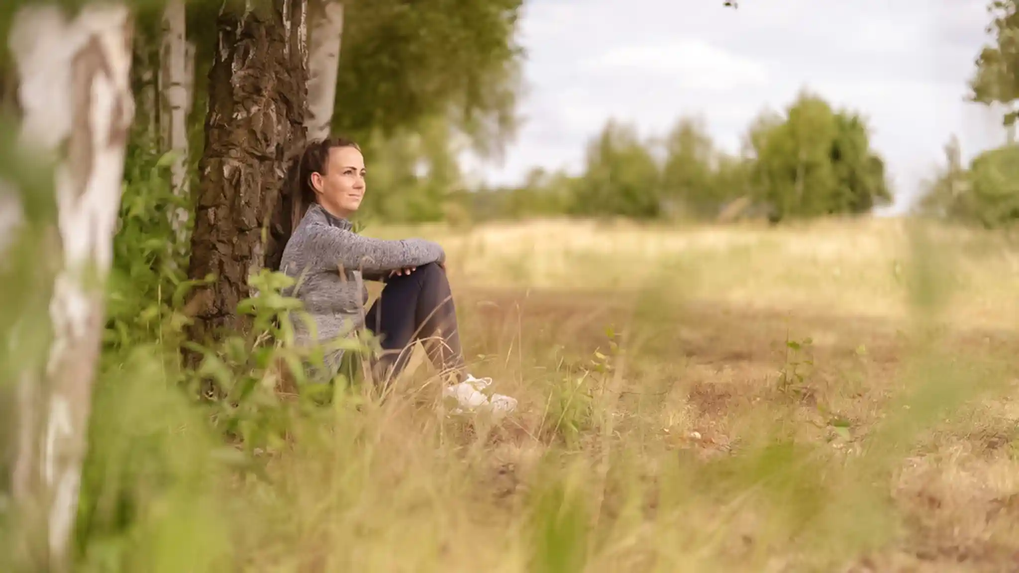 Eine junge Frau sitzt auf dem Boden an einen Baum gelehnt und blickt in die Ferne.