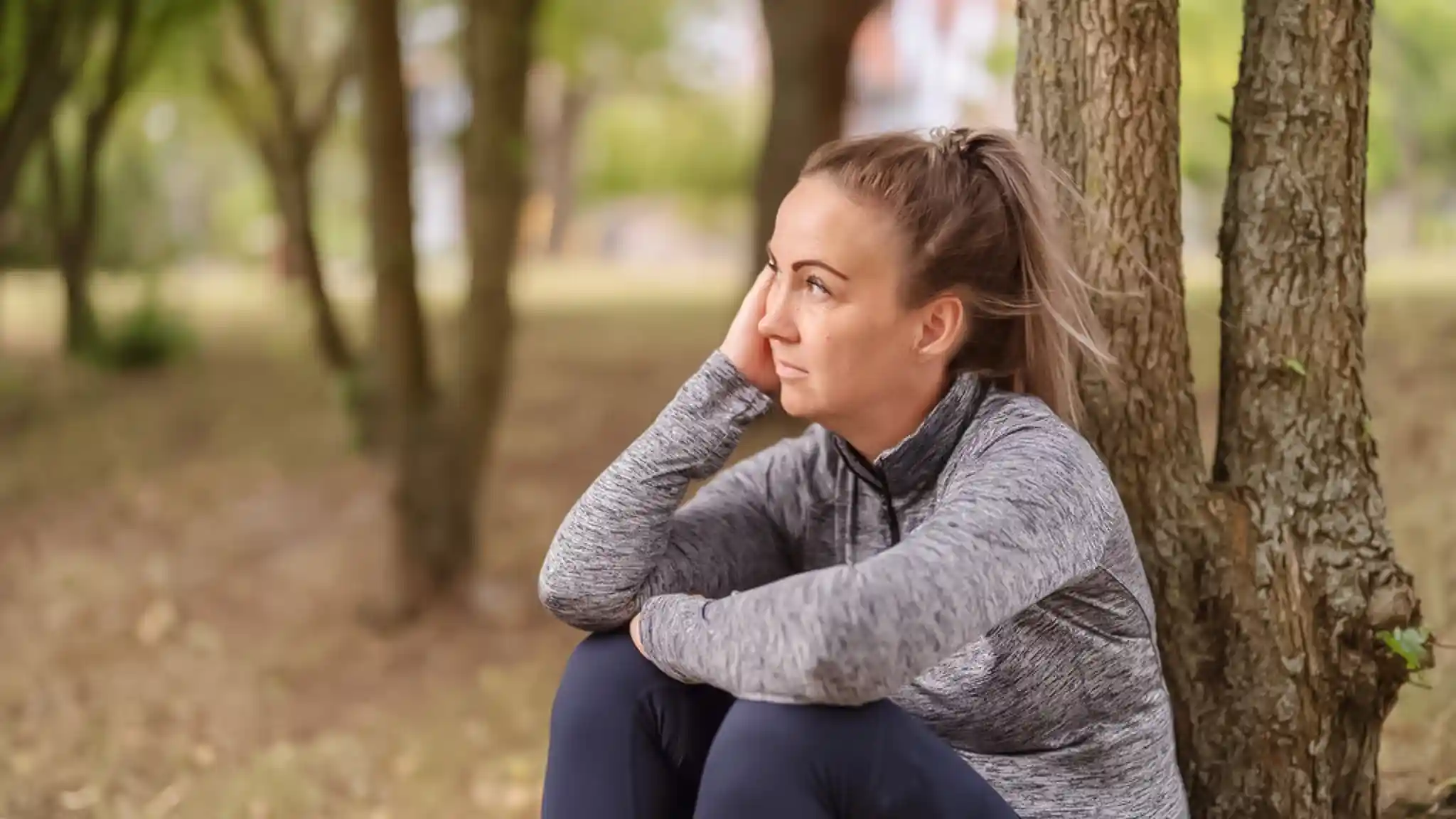 Ein Frau lehnt sitzend an einem Baum und stützt ihr Gesicht auf ihrer Hand ab.