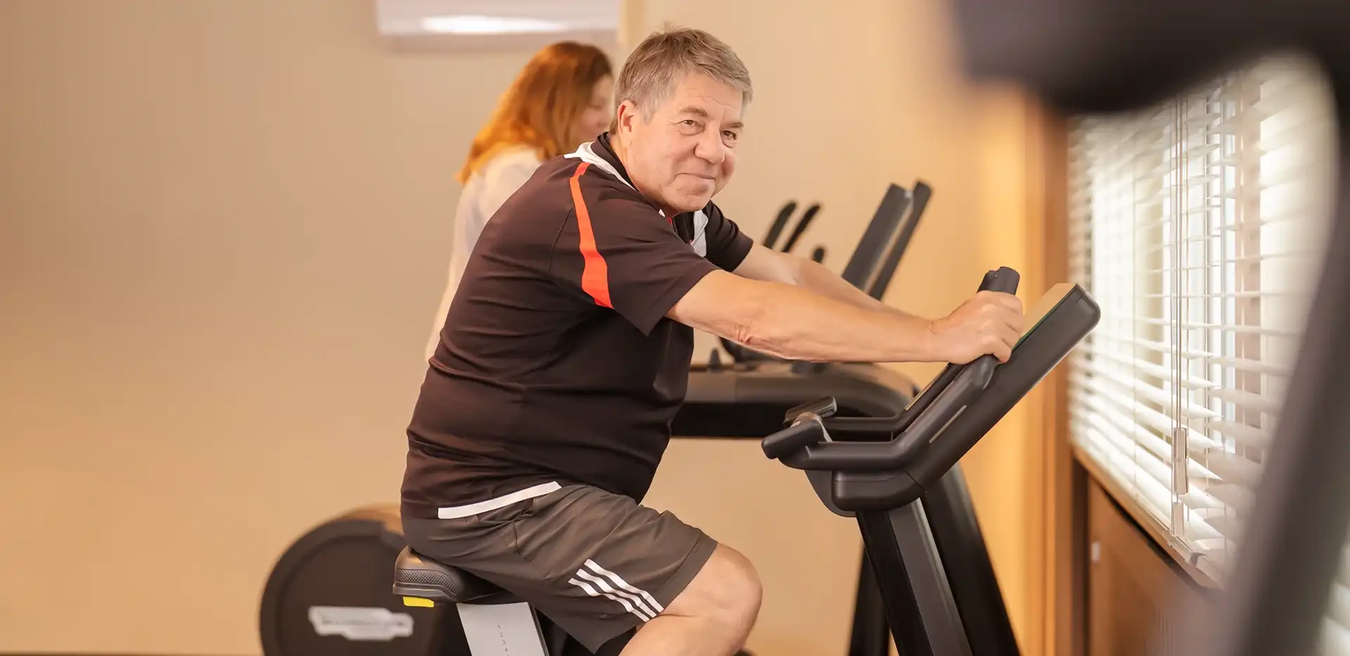 Ein Mann sitzt auf einem Trainingsrad im Fitnessstudio und lächelt in die Kamera.