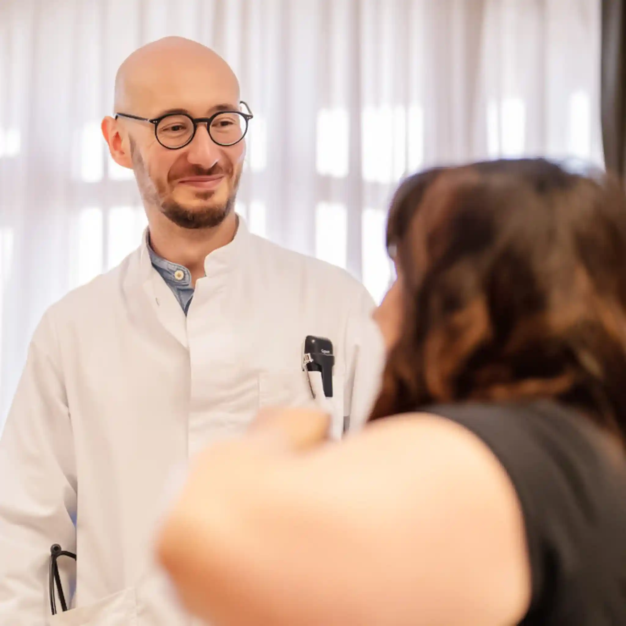 Ein Arzt in weißem Kittel steht neben seiner Patientin und lächelt sie freundlich an.