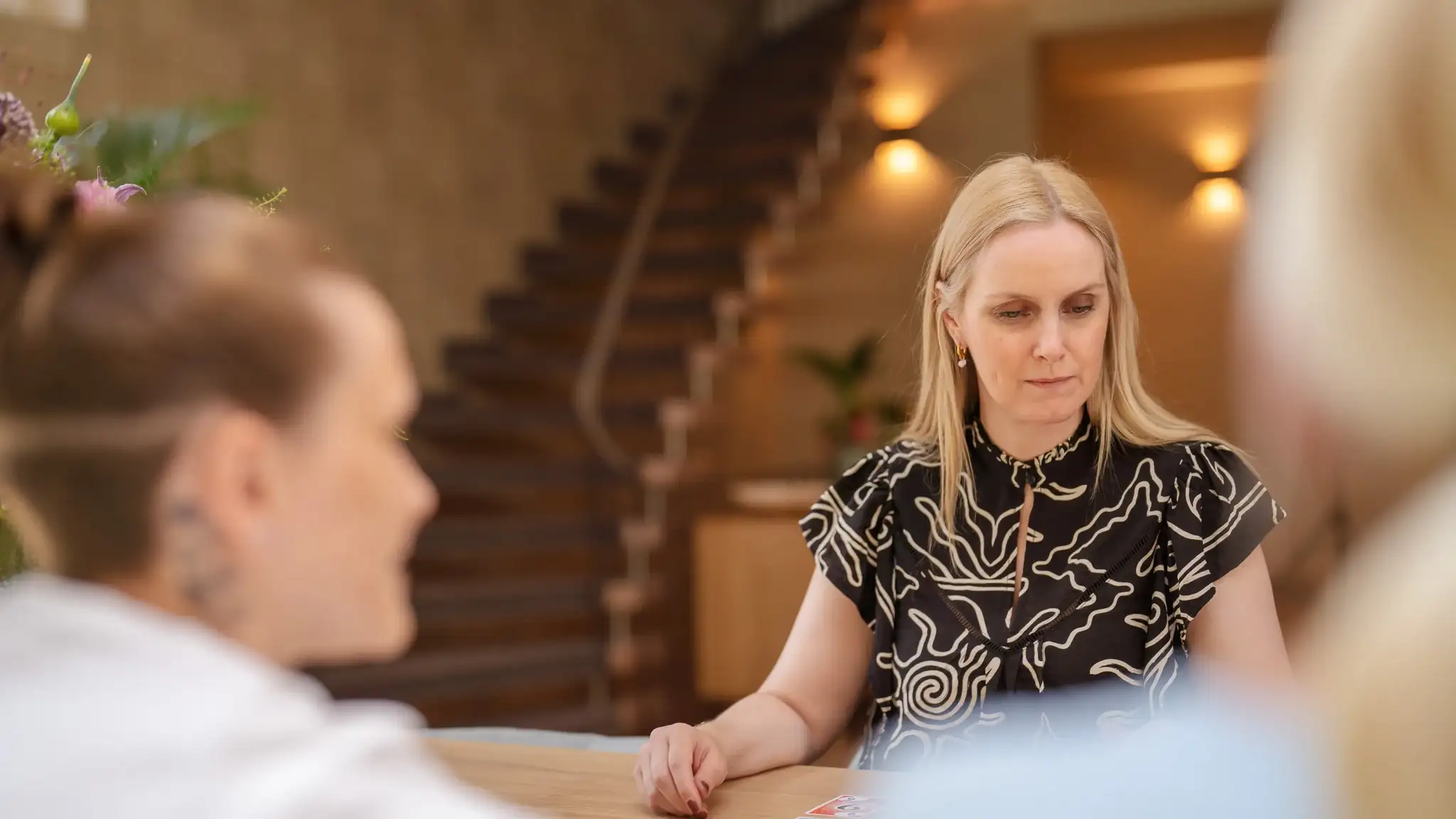 Mehrere Personen sitzen an einem Tisch und spielen Karten. Der Fokus liegt auf einer blonden Frau.