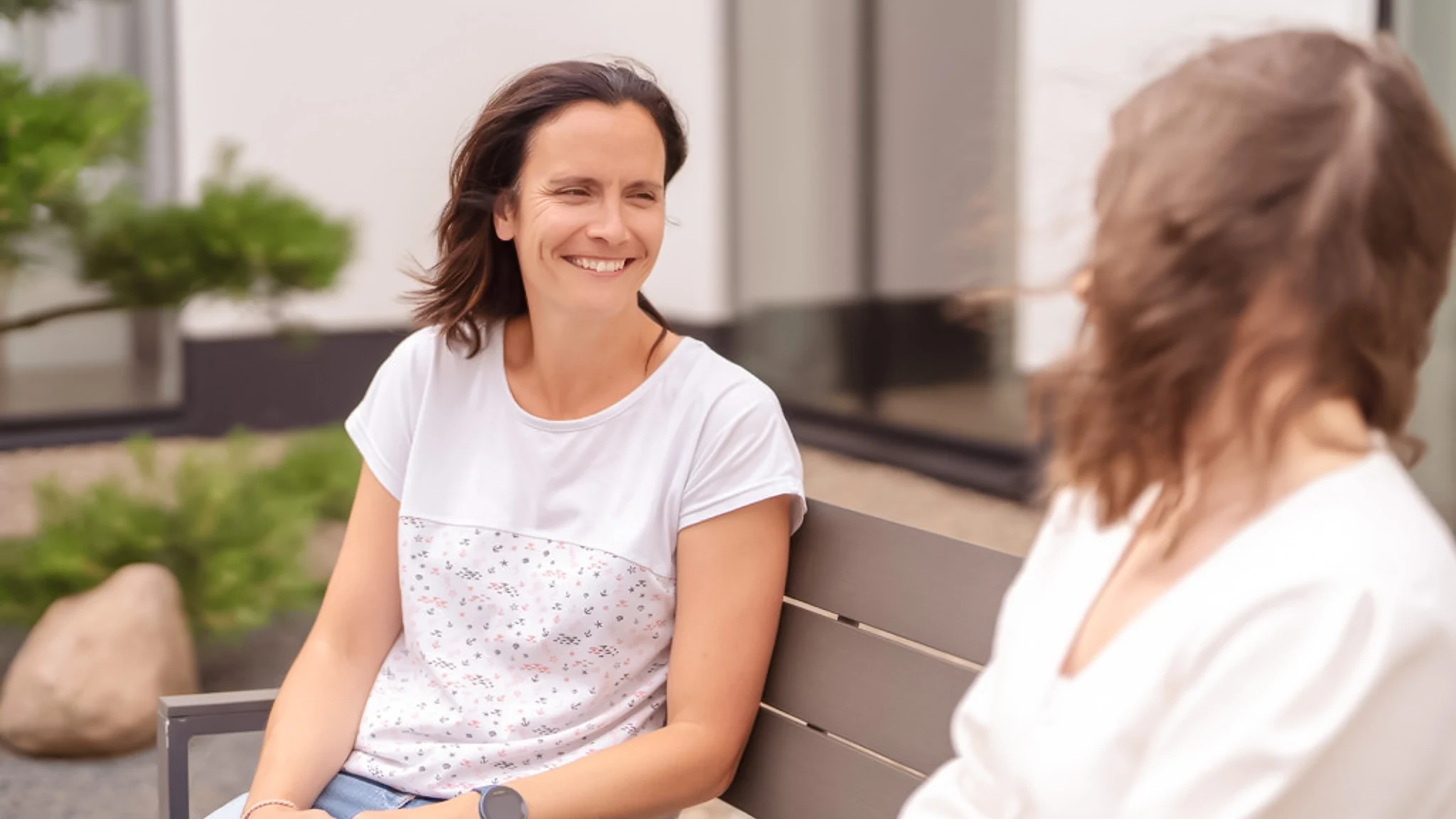 Zwei Frauen sitzen auf einer Bank, die eine blickt die andere lachend an.