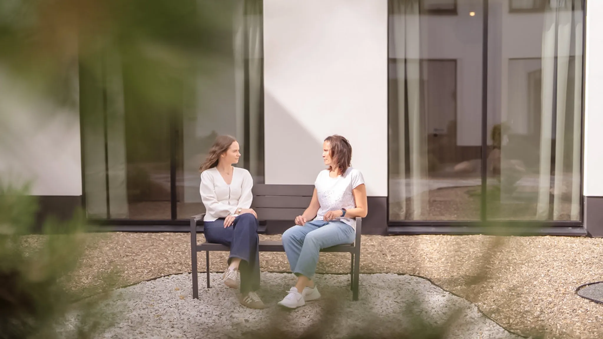 Zwei Frauen sitzen jeweils in einer der Ecken auf einer Parkbank uns schauen sich an.