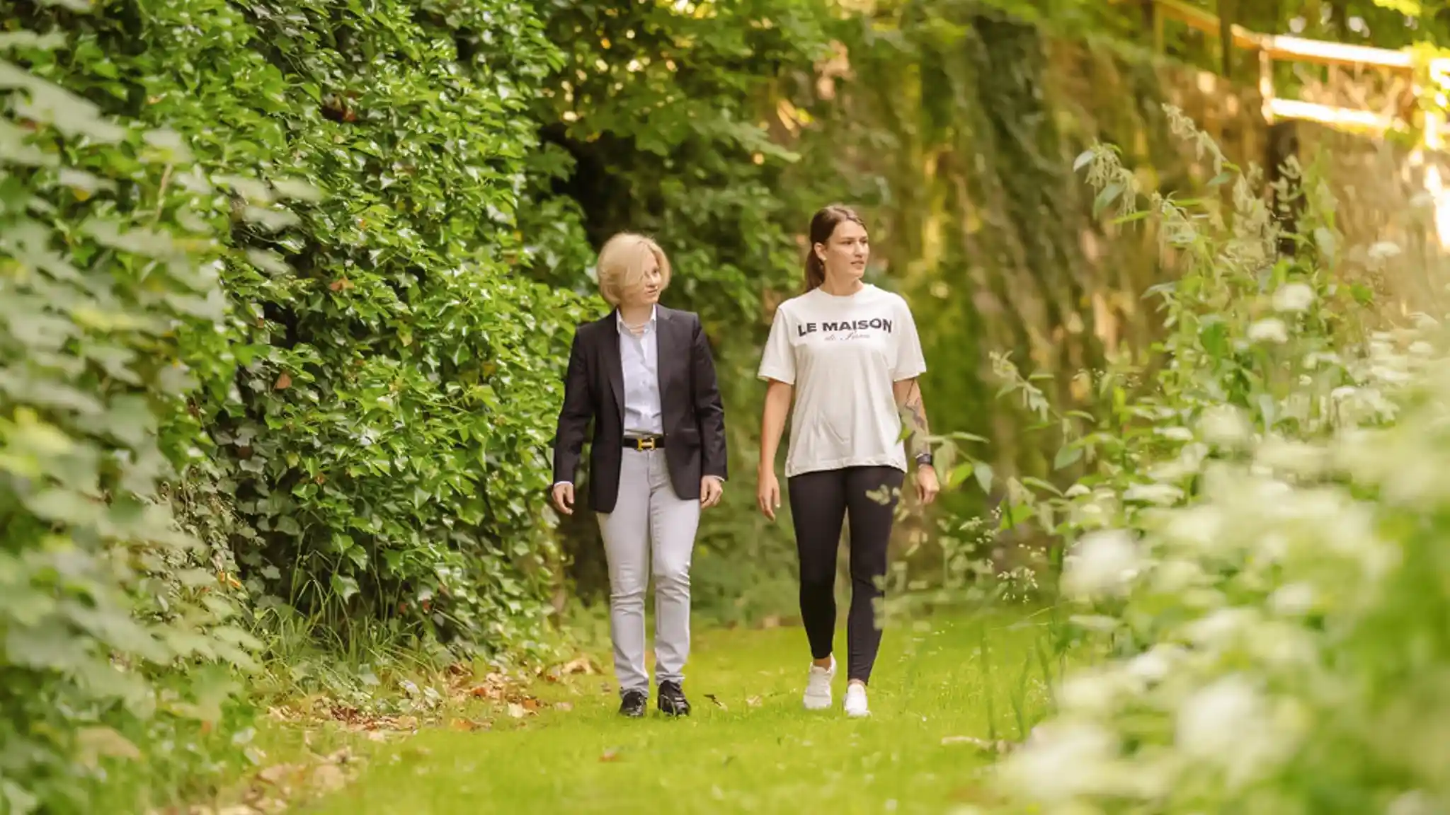 Eine Patientin und eine Ärztin laufen nebeneinander über eine grüne Wiese.