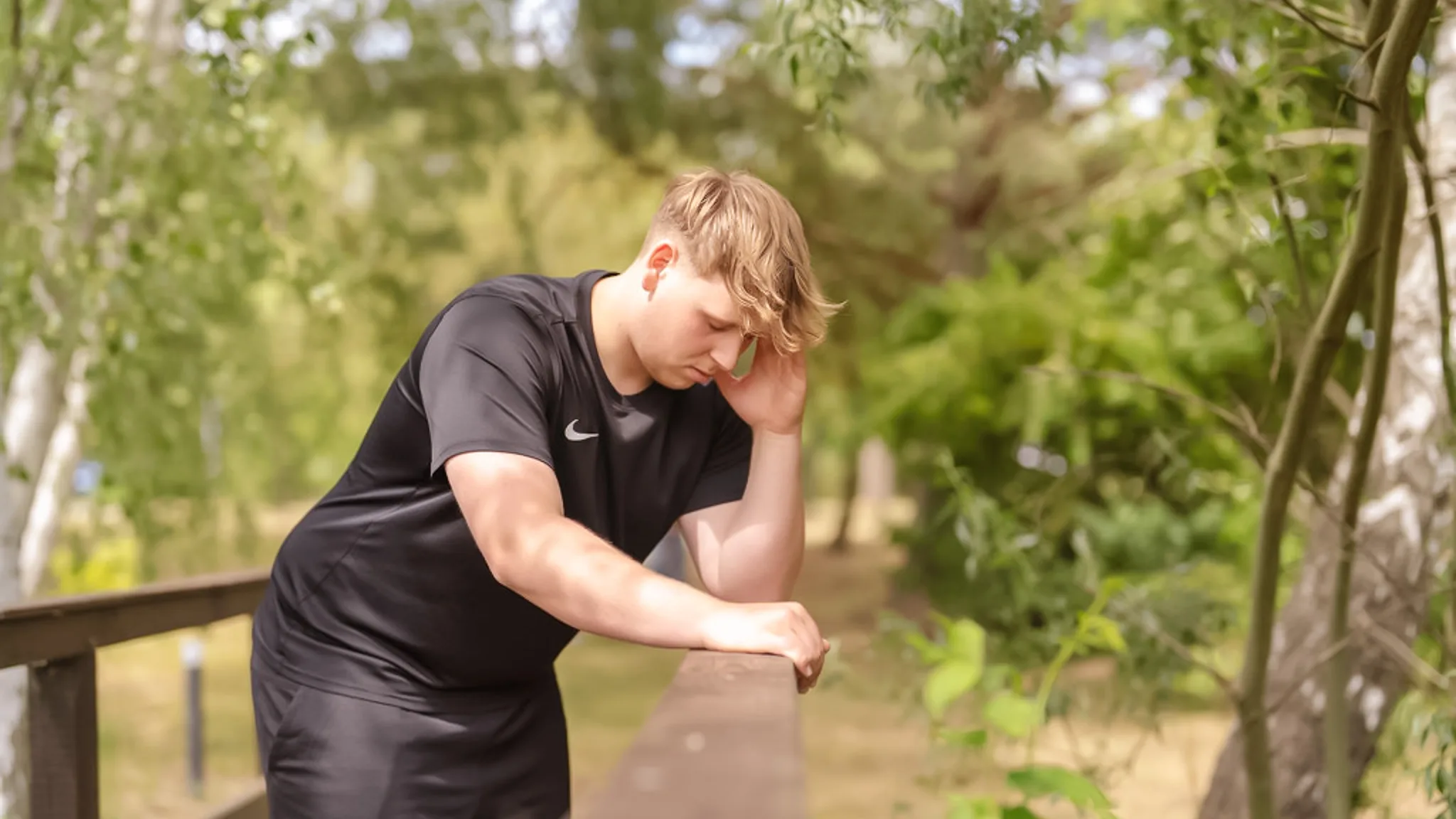 Ein Mann in steht auf einer Brücke im Wald und lehnt mit einen quälenden Gesichtsausdruck am Geländer.