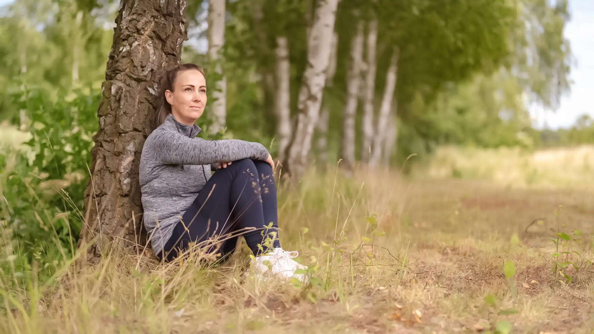 Eine Patientin sitzt auf dem Boden an einen Baum gelehnt und blickt nachdenklich in die Ferne.