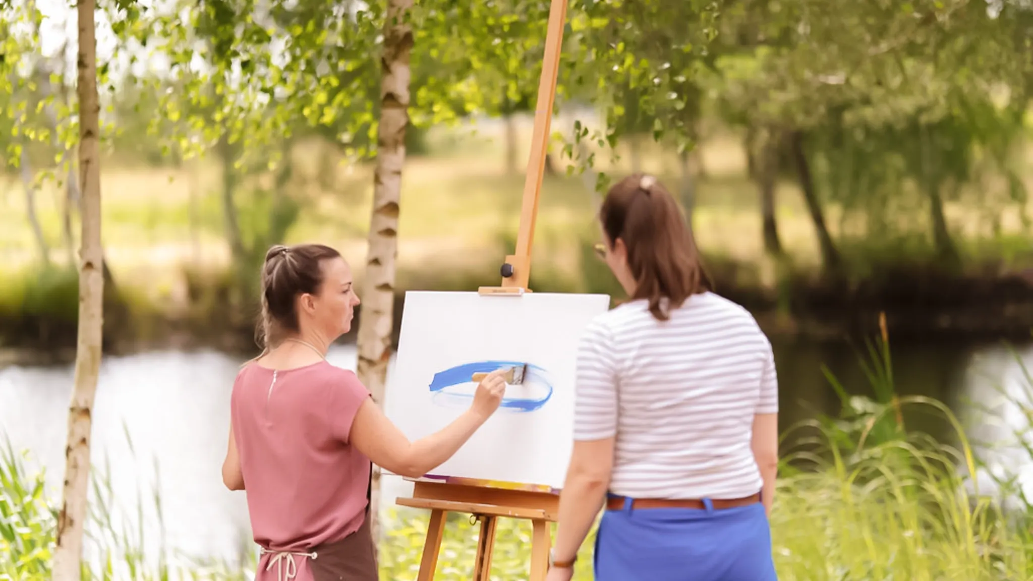 Eine Patientin malt in der Natur auf einer Leinwand. Ihre Therapeutin steht daneben.