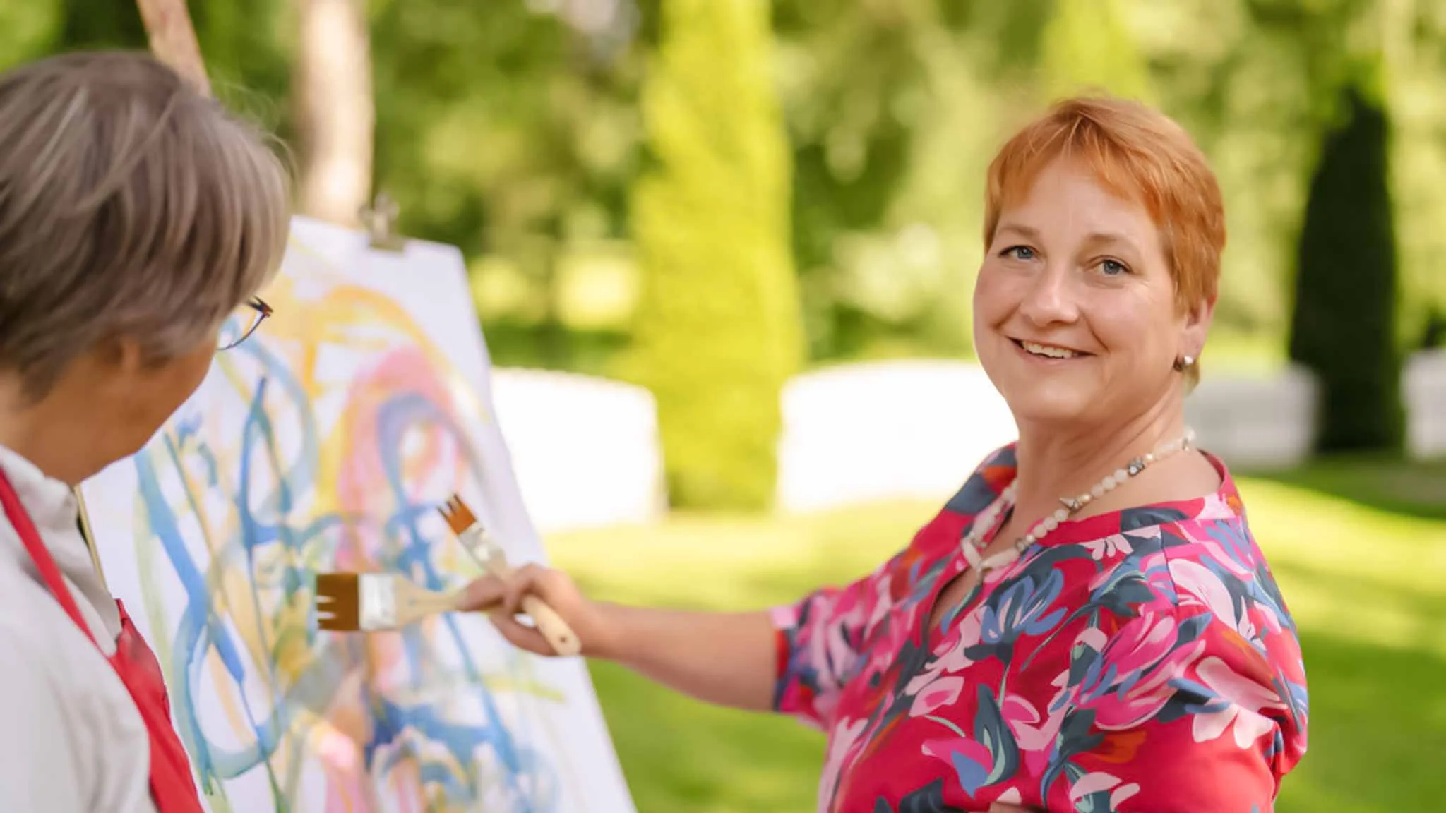Eine Frau in den Wechseljahren mit Depressionen nimmt an der Kunsttherapie teil und malt ein Bild auf einer Leinwand. Sie steht dabei im Garten. 