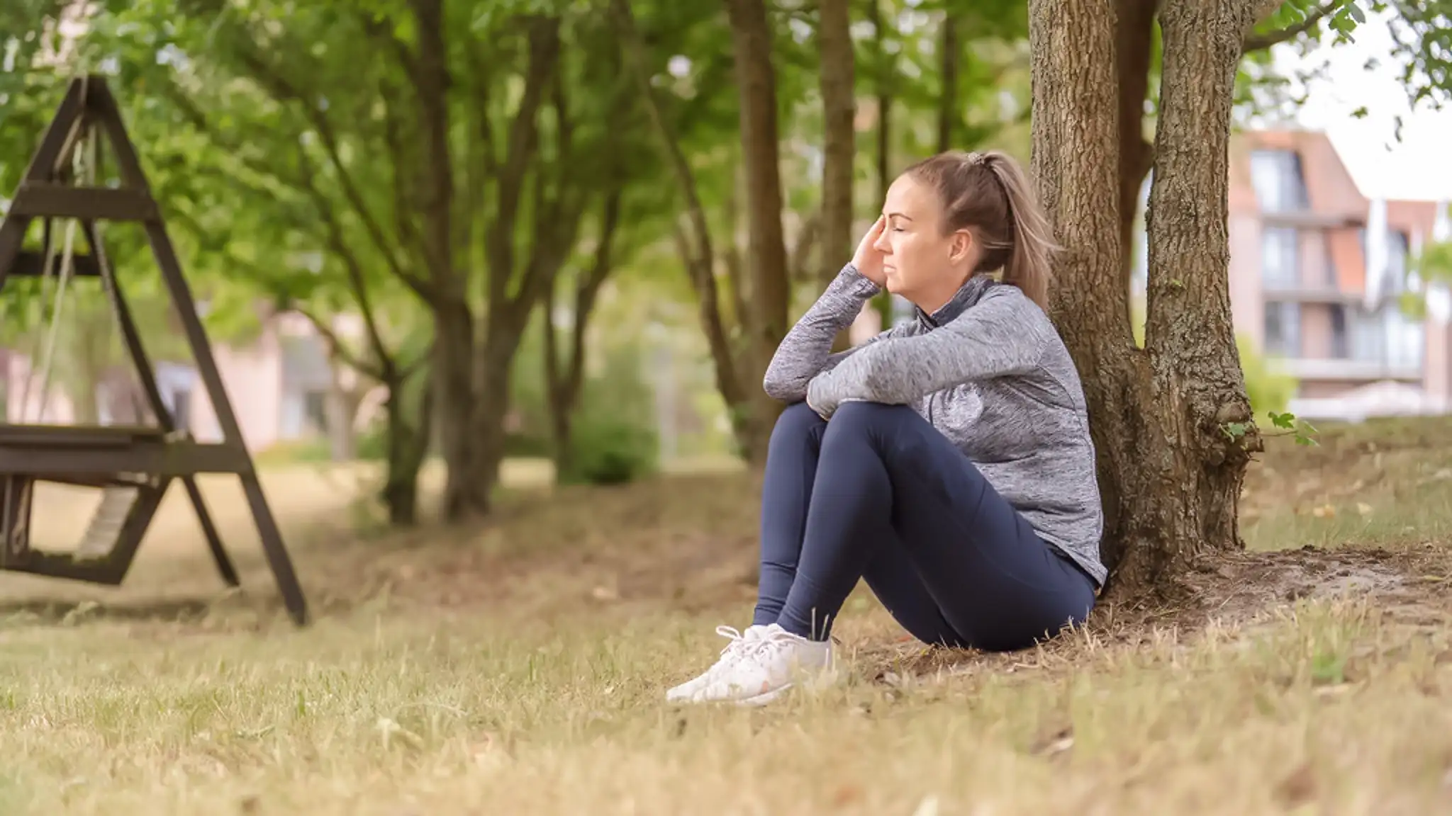 Eine unter Depressionen leidende Patientin sitzt an einen Baum gelehnt auf dem Boden und hat die Augen geschlossen.