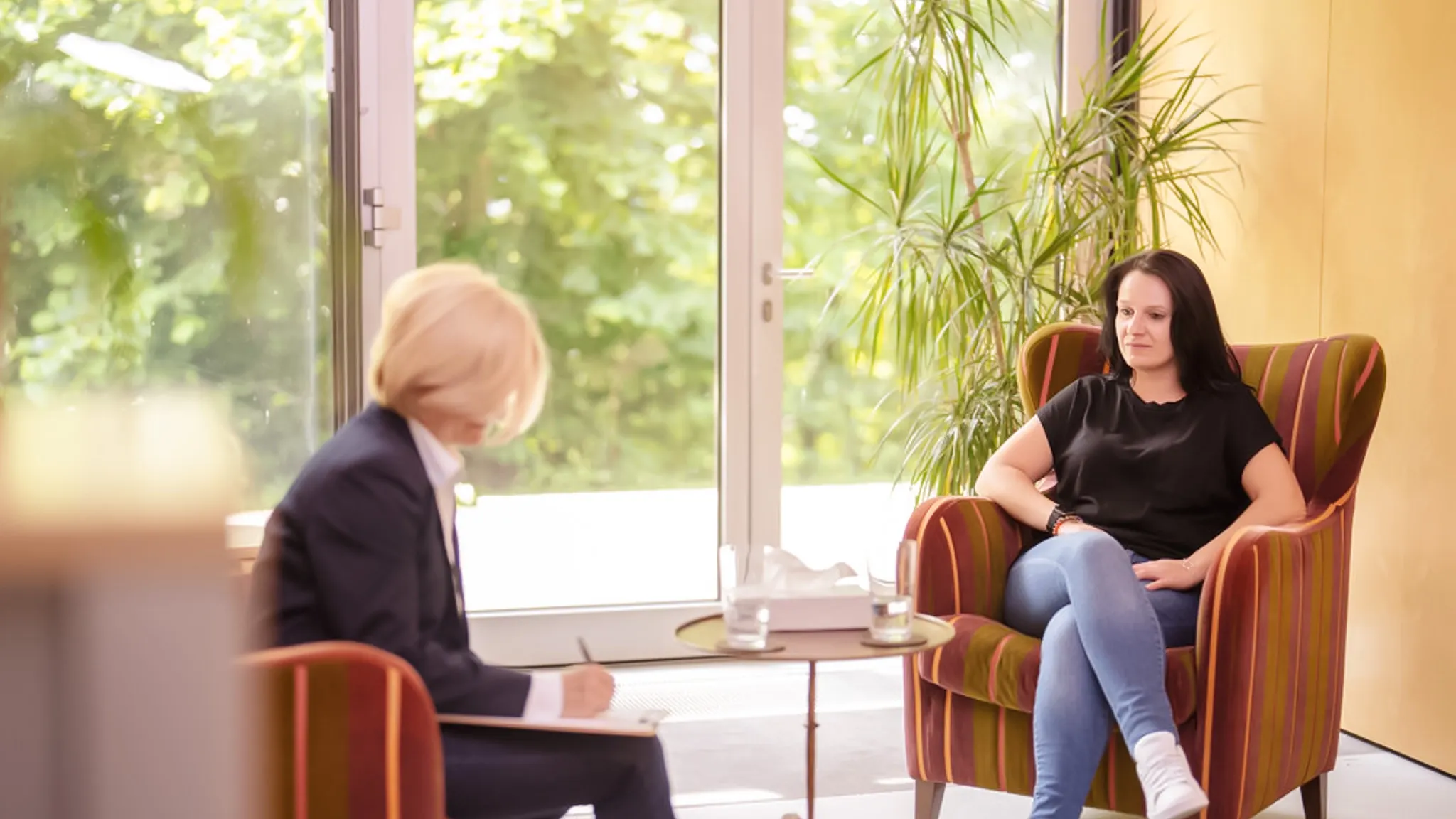 Eine Frau, die unter den Auswirkungen der Wechseljahre leidet, sitzt in einem Sessel. Ihr gegenüber sitzt eine Psychiaterin, die sich Notizen macht.