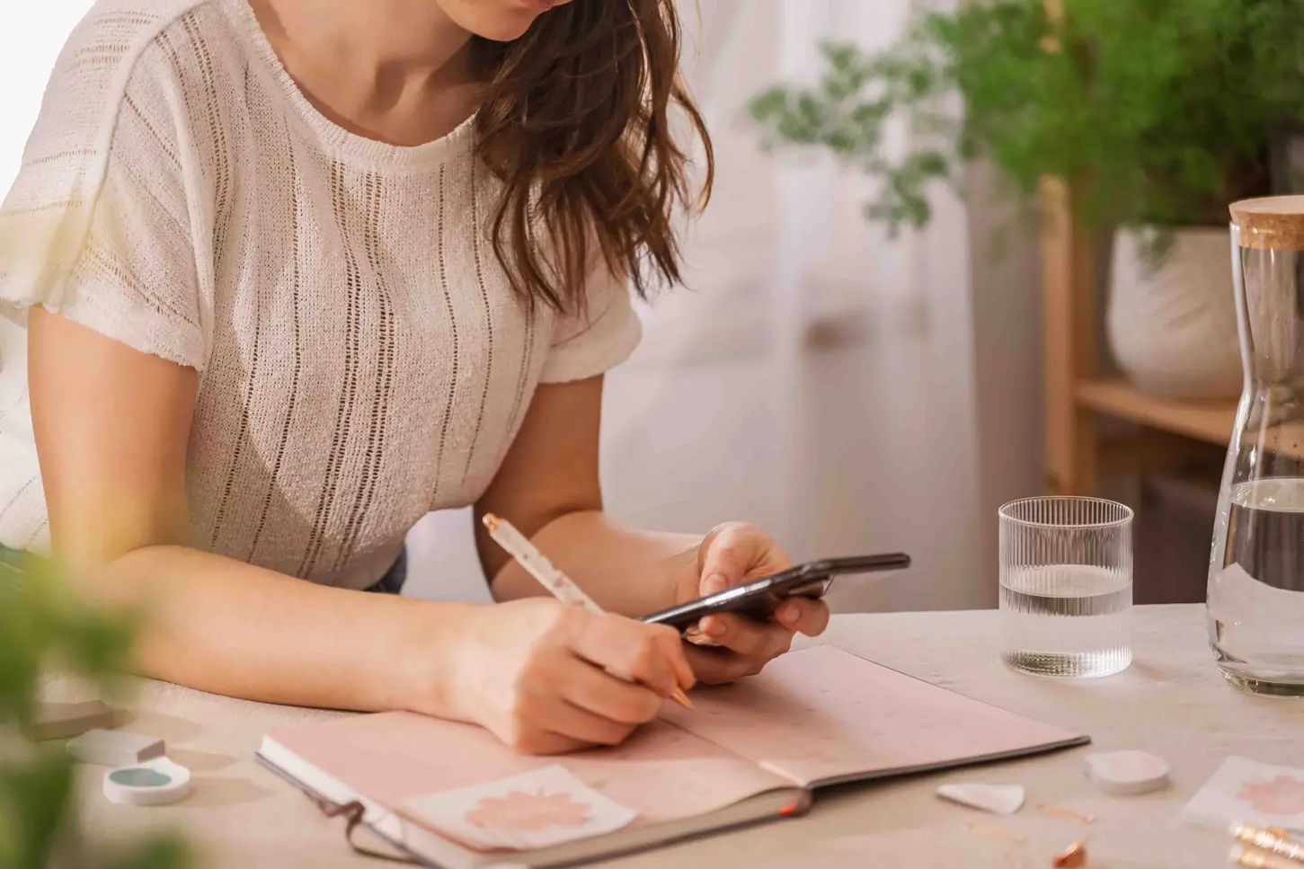 Eine Frau mit Mental Load sutzt an ihrem Handy am Schreibtisch vor ihrem Notizbuch.