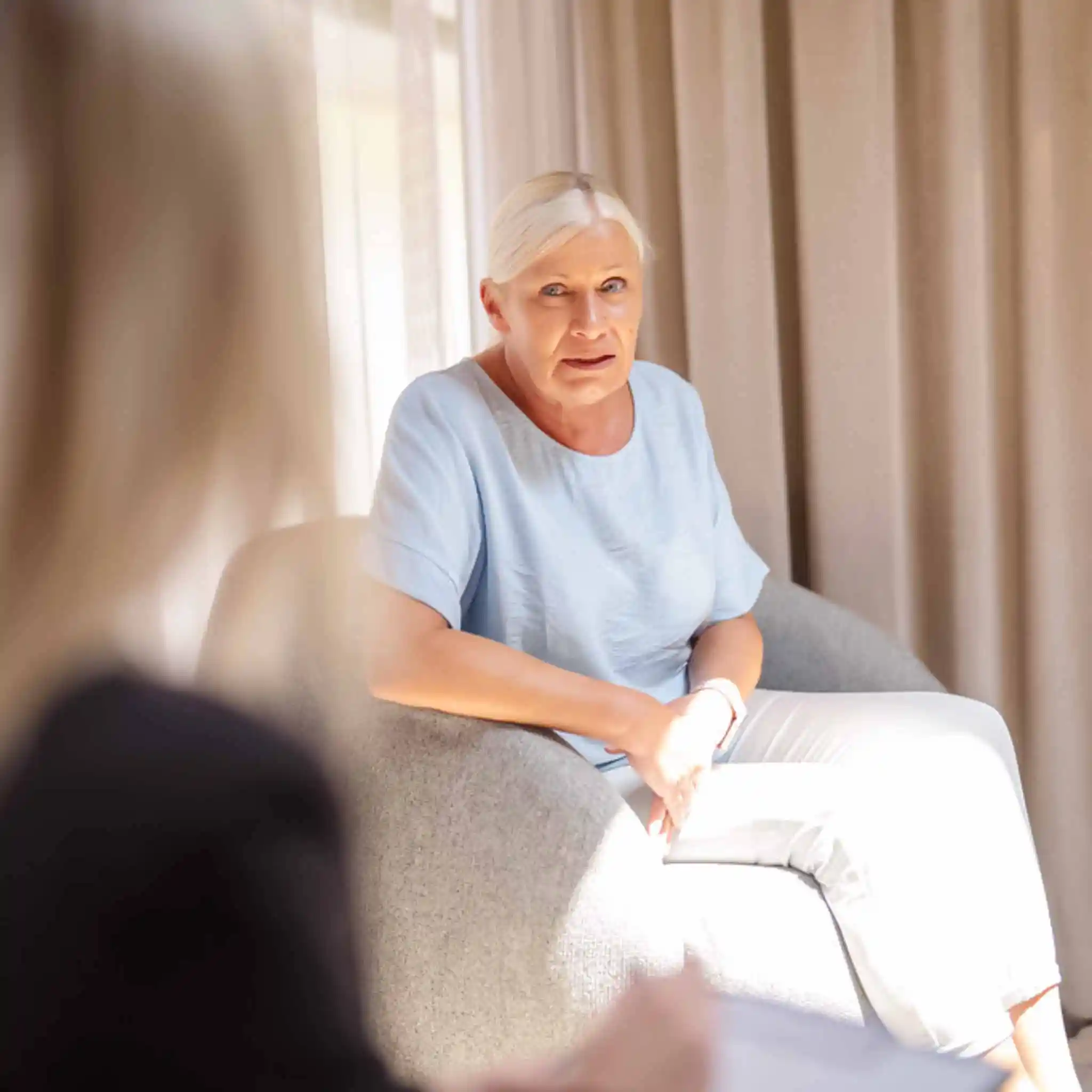 Eine Patientin spricht mit einer Therapeutin während einer Sitzung, Sie sitzt in einem grauen Sessel