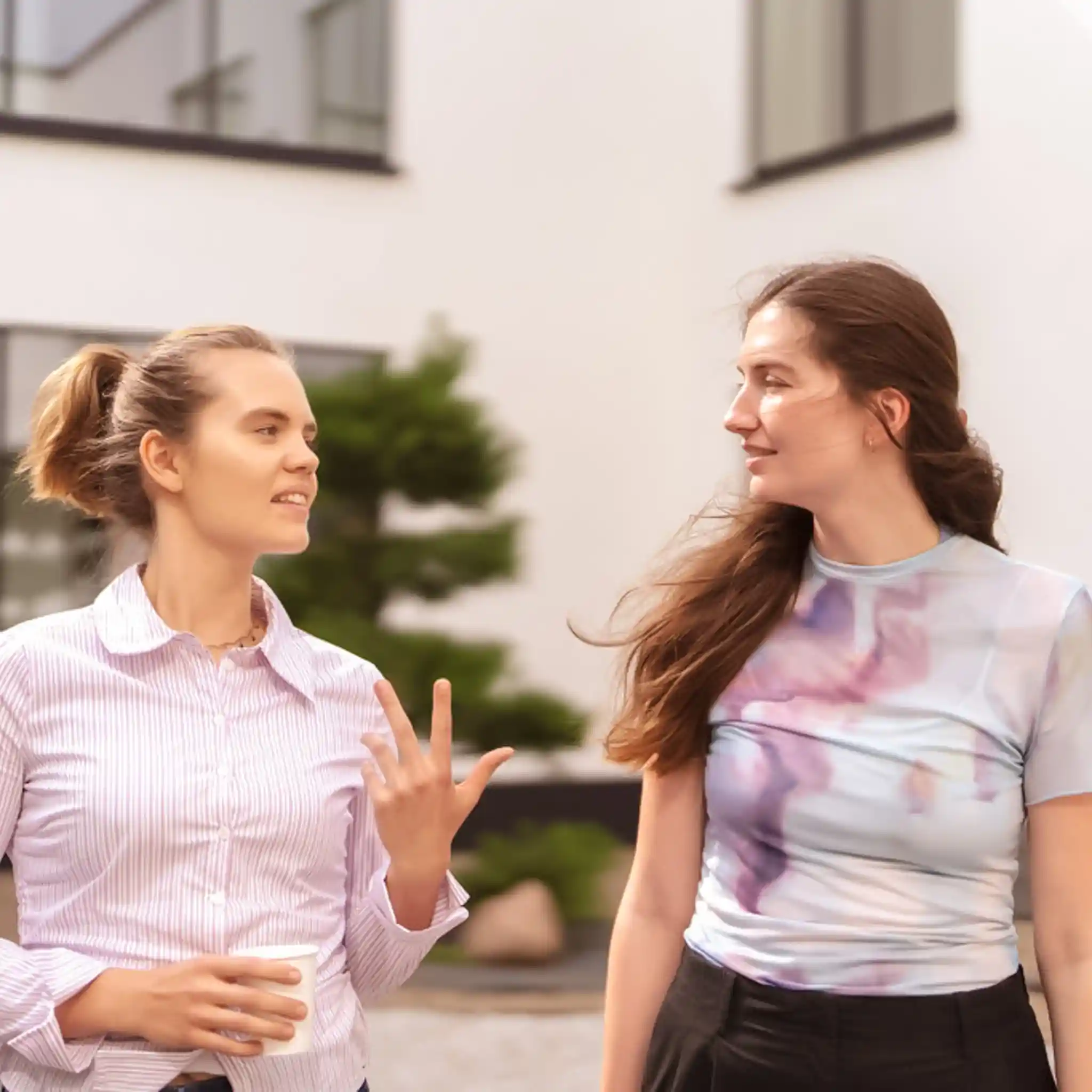 Zwei junge Frauen unterhalten sich im Gehen im Freien, während eine von ihnen einen Kaffeebecher in der Hand hält.