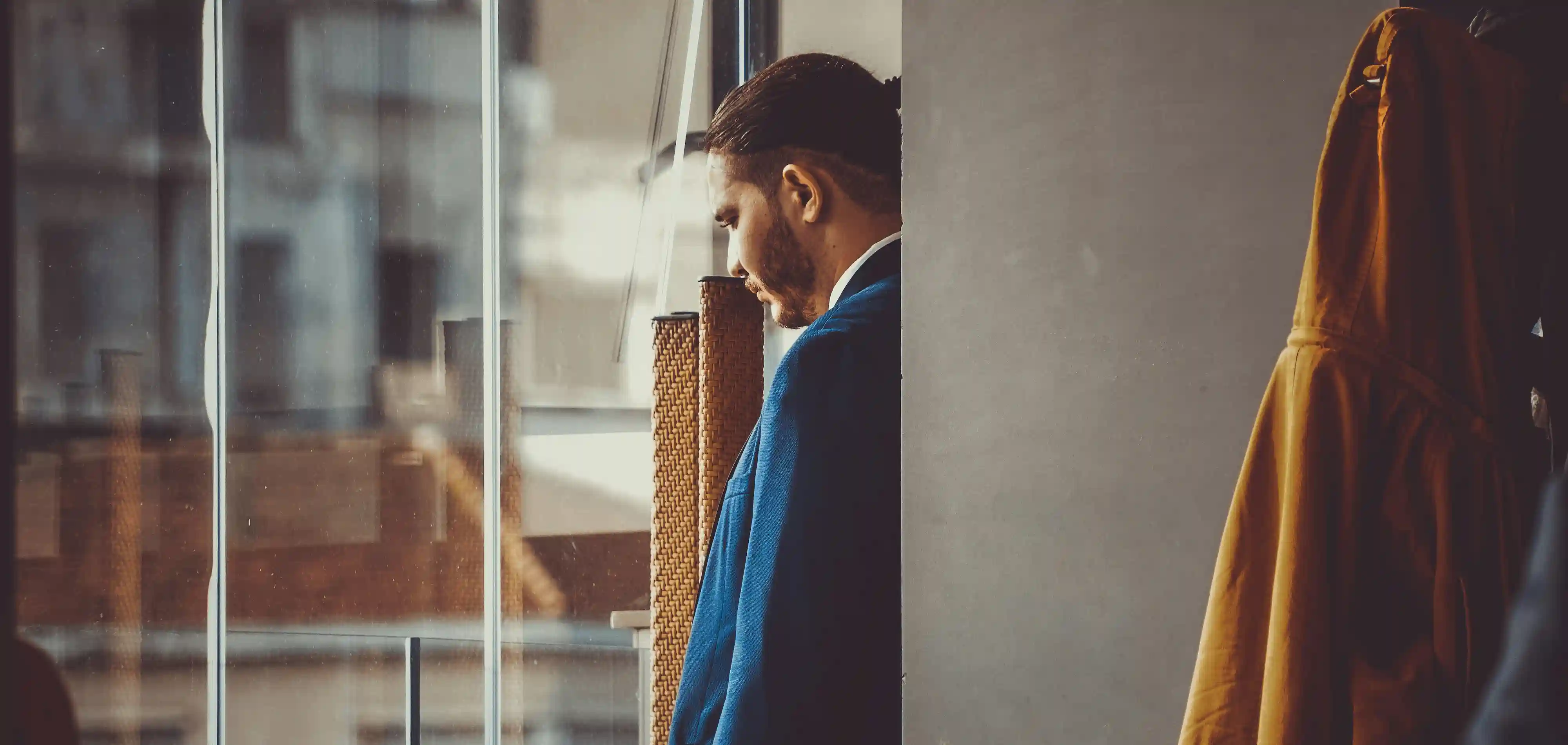 Ein Mann im blauen Sakko steht nachdenklich mit gesenktem Blick am Fenster eines modernen Gebäudes.
